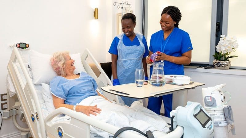 An older woman recovering from surgery at North Bristol Private Hospital. She is smiling up at two bedside nurses.